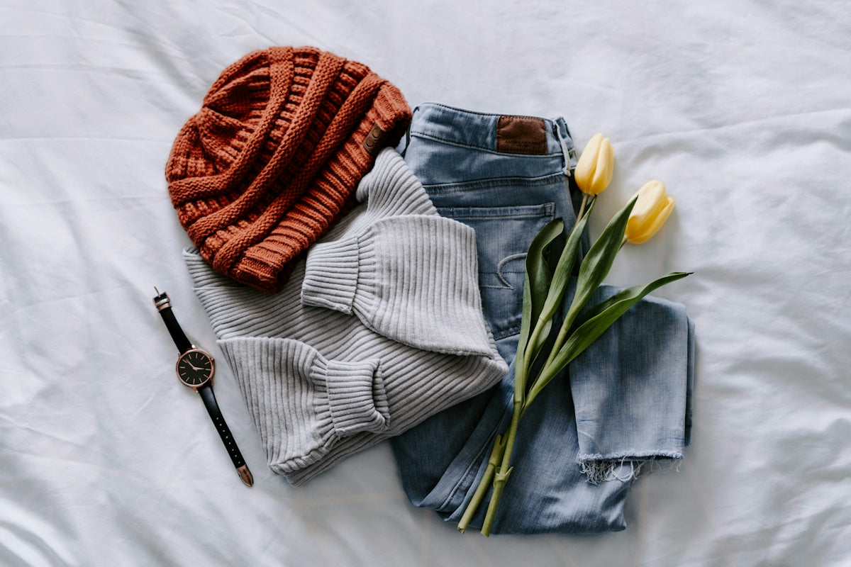 A rust-colored beanie, grey sweater, blue jeans, wristwatch, and yellow tulips on white bedding